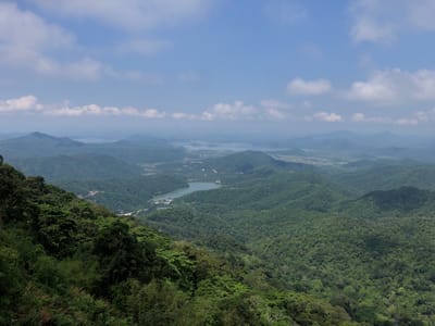 This is a wide-angle, high-angle shot of a vast, green, mountainous landscape under a partly cloudy blue sky. In the foreground, on the left, is a dense area of lush green trees and foliage, showing a steep, downward slope. As the view recedes into the distance, the mountains become less distinct, taking on a hazy, blue-green hue. Several bodies of water, appearing as lakes or inlets, are visible within the valleys and among the hills. Roads and some structures are faintly discernible in the middle ground. The overall impression is one of natural, undeveloped mountainous terrain, possibly a national park or a scenic overlook. The location is identified as Nam Hải, Vietnam, suggesting this is a view from a high point within or overlooking that region. There are no people or animals visible in the image.