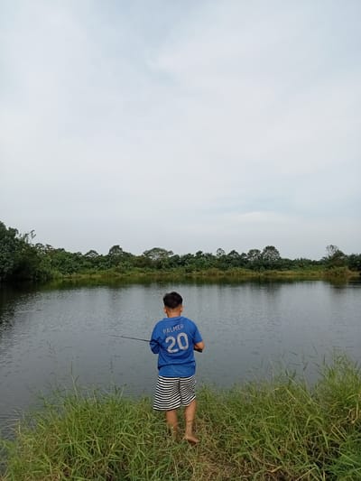 A young boy, facing away from the camera, stands on the grassy bank of a calm body of water. He is fishing, holding a fishing rod. He wears a blue t-shirt with white lettering that reads "PALMER" and the number "20" on the back, and black and white striped shorts. The environment appears to be a natural, rural setting, possibly a lake or pond, surrounded by lush green trees and vegetation. The sky is overcast with a pale, diffused light. The location is the city of Teluk Kenidai, Indonesia.