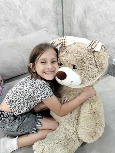 A young girl with brown hair, wearing a leopard-print t-shirt and denim shorts, hugs a large teddy bear while sitting on a sofa. The background consists of a textured grey wall and a light-colored pillow. The scene is indoors.