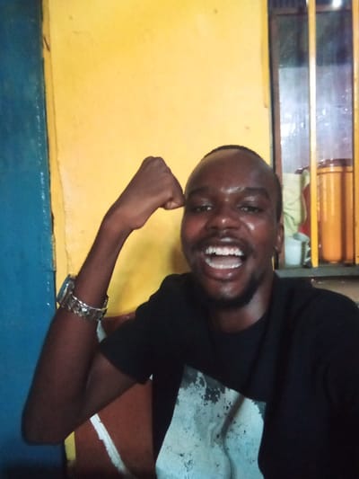 An image taken in an indoor setting within Majengo ward, Kenya, depicts a single male subject positioned centrally. The male, of dark complexion, short dark hair, and a trimmed beard, is wearing a black short-sleeved t-shirt featuring a light-toned, abstract graphic on the chest. His right arm is bent at the elbow, with the hand fisted in a flexing gesture. On his left wrist, he wears a metallic watch and multiple metallic bangles or bracelets. He is smiling broadly with an open mouth, showing his teeth, and looking directly towards the camera. The background consists primarily of a bright yellow wall. To the far left, a vertical section of a blue painted surface, possibly a door frame, is visible. To the upper right, a window with dark, vertical security bars is present. Through or near the window, two cylindrical, yellow-orange containers and one white mug are discernible.