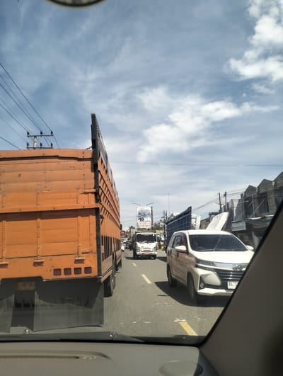 The image shows a view from inside a vehicle, looking out at a busy road in Grong-Grong, Indonesia. A large orange truck is in the foreground, partially obscuring the view of traffic ahead. To the right, a white car is visible in the lane. In the distance, other vehicles and buildings line the road. The sky is partly cloudy. The scene depicts a typical traffic situation on a sunny day.