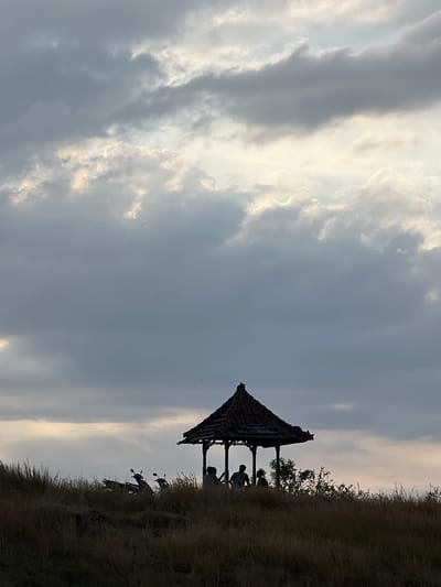 The image shows a silhouette of a small open-air pavilion with a tiled roof, situated on a grassy hill. Three people are visible inside the pavilion, appearing as dark figures against the lighter sky. Two motorcycles are parked on the left side of the hill, also silhouetted. The sky is filled with dramatic clouds, with patches of sunlight breaking through, suggesting either sunrise or sunset. The foreground is dominated by dry, tall grass. The scene depicts a tranquil, natural setting, likely in a rural or semi-rural area, consistent with the landscape around Kubutambahan, Indonesia.