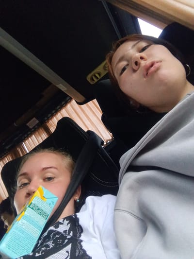 Two young women are in the interior of a bus, with one woman in the foreground holding a juice box up to her face. The woman in the foreground has blonde hair and is wearing a white shirt with black lace detailing and a seatbelt. The second woman, to the right and slightly behind, has reddish-brown hair and is wearing a light grey hooded sweatshirt. The bus interior features dark seats, beige curtains, and overhead compartments, suggesting a travel setting. The scene is captured from a selfie perspective, with the camera tilted downwards. No specific details are visible that would indicate the location in the city of Bol'shoi Bereznik, Russia.