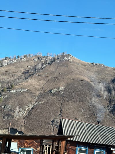 The image shows a mountainside in Manzherok, Russia, with a partly wooded slope and rocky outcrops. Power lines traverse the left side of the frame against a clear blue sky. A corrugated roof casts shadows, and a section of a rustic wooden building with blue window frames is visible on the right. The hillside appears dry, with brown vegetation covering the ground.