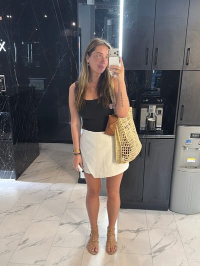 A woman stands in a brightly lit, modern room, taking a selfie in a mirror. She is wearing a black tank top, a white wrap skirt, and gold studded gladiator sandals. She has long, wavy brown hair and is wearing gold hoop earrings. A woven tote bag with a brown leather case attached hangs from her left shoulder. The background features black marble walls, dark cabinets, a coffee machine, and a water cooler. The floor is tiled in a light marble pattern. The location is the city of Укромновское сельское поселение, Ukraine.