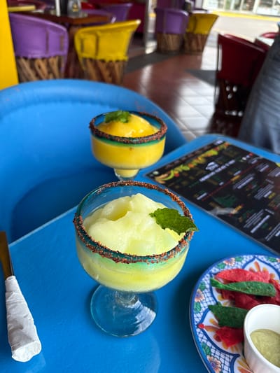 Two yellow frozen cocktails sit on a blue table. One is topped with a mint sprig. Both glasses have a red rim. There is a menu and a small dish with tortilla chips and a cup of guacamole on the table. There are colorful chairs and tables out of focus in the background. The location is a restaurant in Pozos, Costa Rica, at the coordinates 9.951 latitude and -84.194 longitude.