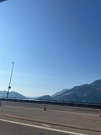 The image displays a daylight scene of an elevated highway segment, identified as the A2, overlooking Lake Lucerne in Beckenried, Switzerland. The asphalt road in the foreground exhibits white solid and dashed lane markings, accompanied by faint red linear markings on the pavement, indicative of recent or ongoing road work. Two red and white striped traffic cones are visible on the road surface: one is centrally positioned within the immediate lane, and another is partially visible at the far right edge of the frame. A dark metal guardrail or barrier runs along the right edge of the roadway, separating it from the large body of water. On the left, a tall, slender street light with a rectangular luminaire head and a yellow square sign attached to its pole extends upwards. Beyond the guardrail, the calm, blue expanse of Lake Lucerne is visible, extending towards a distant backdrop of multiple layers of green, heavily forested mountains, which recede into a light atmospheric haze. The sky overhead is clear and bright blue. No individuals, vehicles, or dynamic actions are depicted within the scene.