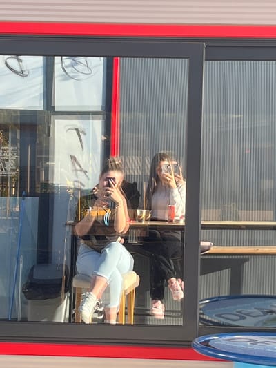 The image is a reflection in a window of what appears to be a commercial space, likely a restaurant or cafe. Two women are visible in the reflection. One woman wears a white shirt and black pants and is looking at her cell phone. The second woman wears a dark-colored shirt and light-colored jeans. She is also holding a cell phone. The reflection shows interior elements such as tables and chairs, as well as signage in the window itself. There is a black suitcase on the floor. The exterior of the building has a red and grey window frame. The setting appears to be Satovcha, Bulgaria.