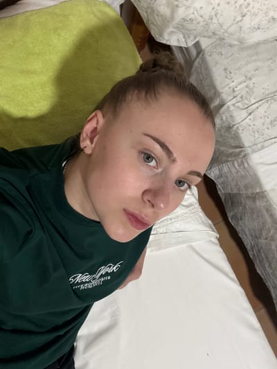The image shows a close-up, high-angle shot of a young woman lying on her side. She has fair skin, light blue eyes, and her blonde hair is pulled back into a bun. The woman is looking up and to her right with a neutral expression. She is wearing a dark green t-shirt with white text on it. The text appears to read "New York Five Boroughs MCMXXII".
The woman is lying on what appears to be a bed with white sheets. To her left, there is a bright green towel or blanket. In the background, there are white sheets with a subtle floral pattern. The lighting suggests it may be indoors, possibly in the evening or with artificial lighting.
The context provided, "Cañada De La Plata, Spain", indicates the location. The scene is intimate and personal, likely a selfie taken by the woman herself. There are no other people or significant objects in the frame. The overall mood is calm and relaxed.