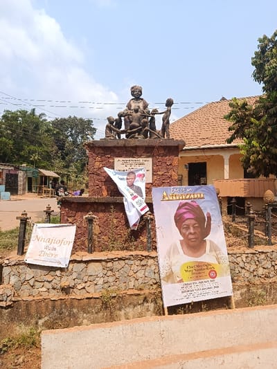 This image depicts a public square or intersection in Ndiaga-Akpugu, Nigeria, dominated by a central monument under a partly cloudy sky. The primary subject is a reddish-brown stone monument with a rough texture, topped by a dark-colored statue. The statue portrays a central adult female figure in traditional attire, seated or standing, surrounded by five children in various poses, suggesting a maternal or community theme. A white plaque with dark text is affixed to the front of the monument's base.

The monument is encircled by a low stone wall, similar in material to the base, with dark metal posts at regular intervals. Attached to these posts and resting on the ground are several banners. On the right, a large vertical banner titled "Exit of an Amazon" displays a photograph of an elderly woman wearing a purple head wrap and a lace top. Text on this banner indicates "Chief Mrs. Ogba Mary Nnaku-Edeh (Nee Nnaji)," "Died 8th January, 2023," and "Burial Saturday 14th January, 2023." To the left, a smaller white banner reads "Nnajjiofor Weds Ebere." A third, partially obscured banner showing a man's portrait is draped over the monument's base.

In the background to the right of the monument, a building with a reddish-brown tiled roof and light-colored walls is visible, featuring multiple windows or openings. A large green tree stands to the far right. To the left, an unpaved road or dirt area extends into the distance, flanked by small, single-story structures with corrugated metal roofs. A solitary individual, dressed in dark clothing, is visible lying or seated low to the ground near these structures. Utility wires traverse the upper left portion of the frame. The overall environment appears dry with sparse vegetation around the monument's base.