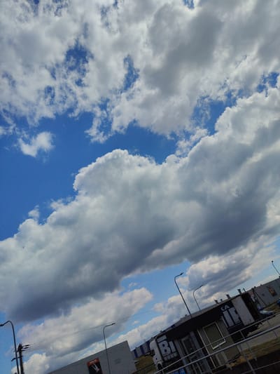 The image shows a cloudy sky over a commercial area. The sky is a mix of blue sky and large, fluffy white clouds, with some darker grey clouds also present. In the foreground, there is a street with a light pole and electrical infrastructure on the left. To the right, there is a low-rise building with a parked truck in front of it. The truck is white and has large dark lettering on its side, which appears to be a logo or advertisement. The building itself is grey and has a fence in front of it. Further in the background, more industrial-looking buildings are visible, along with some parked cars and trees. The overall impression is of a partly cloudy day in an urban or suburban setting. The location is identified as Lomna-Las, Poland.