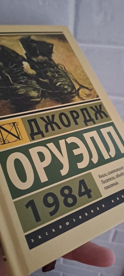 This is a close-up, angled shot of the cover of a book titled "1984" by George Orwell. The cover features a dark, somewhat abstract illustration on the left, and bold text on the right. The title "1984" is prominently displayed in large, dark numerals within a green rectangular block. To the left of the numbers, the name "ДЖОРДЖ ОРУЭЛЛ" is written vertically in large, white letters against a dark green background. Below this, there is a smaller yellow block with a geometric design. Along the right edge of the book cover, more text is visible vertically in dark letters. Some of this text is partially obscured, but it appears to be in Cyrillic script. The book has a cream-colored cover and appears to be a paperback. The background behind the book is a light-colored, textured surface, possibly a wall or brickwork. The image is well-lit, with no distinct shadows indicating time of day. The focus is on the book cover, with other elements slightly blurred.