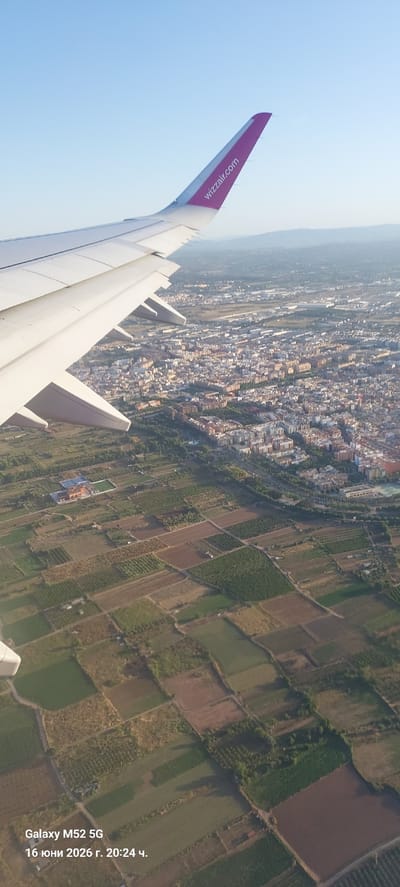 An aerial view shows the wing of an airplane, identified by the "wizzair.com" logo on its purple tip, extending into the frame from the upper left. Below the wing, a sprawling city with a mix of residential and commercial buildings is visible, characterized by numerous houses with reddish-brown roofs and some larger apartment complexes. Agricultural fields, divided into rectangular plots, surround the urban area, with varying shades of green and brown indicating different crops or stages of cultivation. In one of these fields, a complex with a light-colored roof and a green rectangular area, possibly a sports field, can be observed. The environment suggests a daytime scene with clear skies. The image includes a timestamp in the lower left corner reading "Galaxy M52 5G 16 юни 2026 г. 20:24 ч.", which translates to "Galaxy M52 5G June 16, 2026 20:24". The city is identified as Quart de Poblet, Spain.