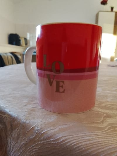 The image displays a two-toned ceramic mug, predominantly red on its upper half and light pink on its lower half, separated by thin horizontal bands of dark red and light pink. The word "LOVE" is inscribed vertically in gold-colored capital letters on the mug's side, which features a white C-shaped handle on the left. The mug rests on a light-colored, patterned tablecloth. The background is out of focus, revealing a dark object on a surface and a striped piece of furniture (possibly a sofa or bed) to the left. To the right, a light-colored cabinet with a small red decorative item on top is visible, illuminated by a strong light source from behind. The scene is situated within a domestic interior in Neunkirchen, Germany.
