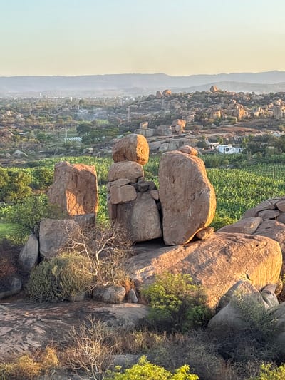 The image shows a landscape in Kamalapura, India, characterized by clusters of large, weathered boulders. The boulders vary in size and shape, and are piled atop one another, creating unusual formations. Vegetation, including green plants, shrubs, and a dry tree, grows around and between the rocks. In the distance, a view opens up to a wider landscape of trees and buildings. The lighting suggests either sunrise or sunset, with soft, diffused light washing over the scene.