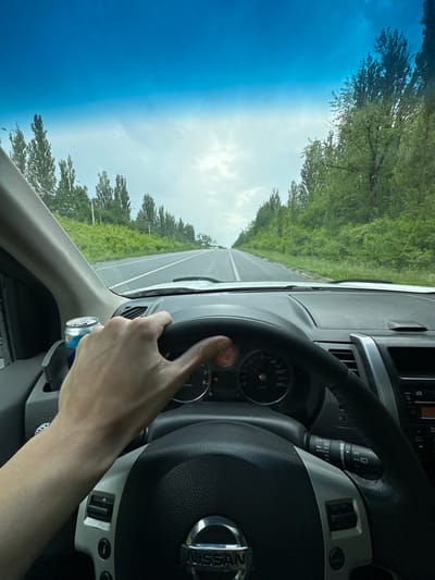The image shows a first-person perspective from inside a car, likely driving on a highway. A hand is visible on the steering wheel, which has a Nissan logo in the center. The dashboard displays a speedometer with readings up to 240 km/h and other indicators. A windshield wiper control stalk is also visible.
Through the windshield and the side window, a bright, sunny sky with some clouds is seen. Lush green trees line both sides of the road, suggesting a rural or suburban setting. The road appears to be asphalt with lane markings. The overall impression is a daytime drive under pleasant weather conditions.
The visible text includes "NISSAN" on the steering wheel, and various labels on the dashboard controls, such as "ACCEL/RES", "LOAD", and "OFF", "AUTO", indicating the car's features. There is also some text on a small object hanging from the dashboard, possibly a car air freshener, which includes "-CARNITINE VITAMIце".
Given the provided location context of Shatava, Ukraine, this image captures a scene of someone driving through a scenic route in the Ukrainian countryside on a clear day.
---
**Note:** The provided OCR output shows some Ukrainian characters and what appears to be a brand name or product description. The description adheres to the factual and concise requirements, incorporating visible text and scene details.