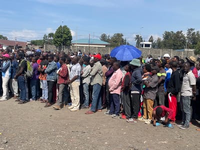 The image shows a large group of people standing in a line. They are all men, and most are wearing casual clothing. There are a few trees and buildings in the background. One man is tying his shoe in the foreground. The scene is likely taking place in a public space, perhaps a street or a market. The image was taken in the city of Ville, Democratic Republic of Congo, at coordinates -1.663 latitude and 29.176 longitude. The people are standing in a line, waiting for something. The men are interacting with each other, speaking, and making eye contact.