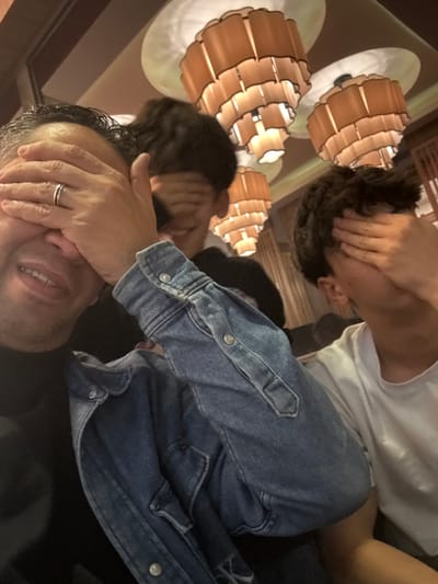 Three people cover their eyes with their hands. The person closest to the camera has dark hair and is wearing a black shirt. There is a silver ring visible on their finger. Behind them is another person with dark hair wearing a denim jacket. The furthest person wears a white t-shirt and has dark hair. Three decorative light fixtures are visible on the ceiling.