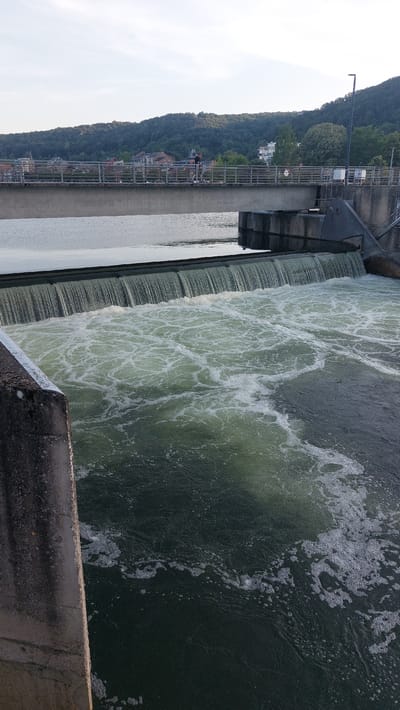 The image shows a dam with water cascading over it. A pedestrian bridge spans the river above the dam, with a person walking across it. The background features a forested hillside with buildings nestled amongst the trees, suggesting an urban or semi-urban environment. The water is turbulent and foamy below the dam, indicating a significant flow. The concrete structure of the dam and bridge is visible, as is the dark water and the lush green hillside. The sky is a pale blue with some clouds. The location appears to be in the city of Namur, Belgium, likely along the Meuse River.