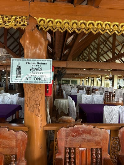 The image shows the interior of a restaurant in Tambon Ko Panyi, Thailand, featuring a rustic wooden structure with ornate golden decorations. A prominent sign is attached to a large wooden pillar, which reads "Please Return Empty Coca-Cola Bottles To Your Dealers AT ONCE!". The restaurant is furnished with numerous tables covered with purple and white lace tablecloths, and chairs draped in white lace covers. Several ceiling fans are visible, along with a red fire extinguisher mounted on the pillar. The overall scene suggests a well-maintained, traditional dining establishment.