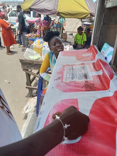 A street vendor in Ovwian, Nigeria, is selling products from behind a large red sign. She is seated in a blue chair, facing forward, and holding a white cloth. She is wearing a yellow and black striped top. A man's arm, wearing a bracelet with multiple silver crosses, reaches up toward the sign from the bottom of the frame. The vendor is sitting in front of a wooden table with various plastic bottles on it, covered by a green plastic cover.  There is a white canopy above the vendor and table. The location is near a busy street, with other people in the background. There is a red car parked in the distance. The ground is paved with concrete.  The vendor appears to be ready to help customers.