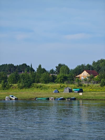 The image depicts a rural landscape, likely near the village of Galanino, Russia, characterized by a wide river in the foreground and a treeline with scattered houses in the background. Several small boats are moored along the grassy riverbank. On the shore, two tents and a dark-colored car covered with a green tarp are visible, suggesting a camping or recreational activity. The sky is a clear blue with a few wispy clouds. The overall scene is serene and suggests a peaceful, natural setting.