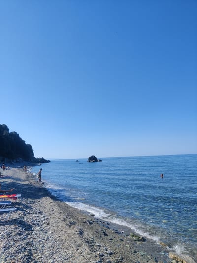 A wide, sunlit shot captures a tranquil pebbled beach and the calm blue sea under a clear, cloudless sky. On the left, a dense, dark green tree-covered hill slopes down towards the water. The beach itself is sparsely populated. In the foreground, lounge chairs with colorful towels are set up. A young boy in green shorts stands at the water's edge, facing away from the camera, appearing to be picking up something from the shallow water. Further along the shore, several adults are visible, some relaxing on lounge chairs and others walking. In the middle ground of the sea, a solitary person swims, their head visible above the water. In the background, two dark rock formations rise from the sea, one significantly larger than the other. The water is clear, allowing the pebbled seabed to be visible near the shore. The overall scene is one of peaceful seaside leisure.