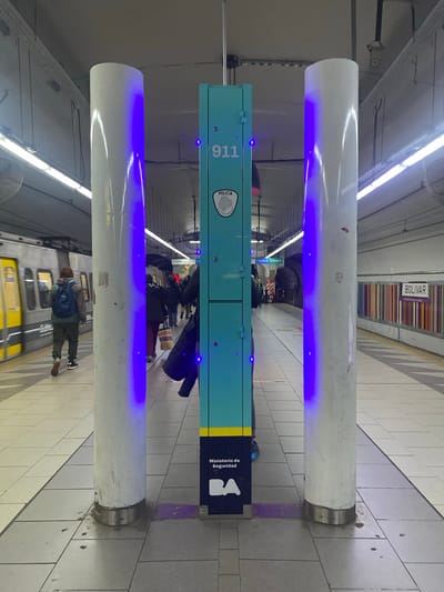 The image shows an indoor scene, most likely a subway station platform in the city of Buenos Aires, Argentina. A blue-green pillar with the number 911 and the Argentine Police logo stands between two white cylindrical structures emitting blue light. People are standing around the pillar, some waiting, and a train appears to be approaching or departing. A sign for the "BOLIVAR" station is visible overhead. The floor is tiled. The scene is well-lit with fluorescent lighting.