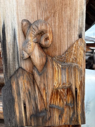 An outdoor, close-up, eye-level shot captures a wooden carving of a ram on a weathered wooden surface. The carving is intricately detailed, depicting the ram's head with large, curved horns, its body, and its legs, with a mountainous background carved behind it. The wood exhibits prominent grain patterns and variations in color, ranging from light tan to dark brown and black, suggesting age and exposure to the elements. There are at least two visible nails securing the carved piece to the wooden background. The background of the image shows a blurred outdoor scene with structures and possibly vegetation. The carving is the primary subject and is centered in the frame.
