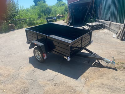 A black utility trailer with a metal frame and black sides sits on a cracked concrete surface. The trailer has a single axle with a silver rim and tire. A red and white sticker is visible on the tire. The trailer has a hitch with a silver coupler and a red handle. A black cable runs from the hitch to the trailer. The trailer is empty. In the background, there is a building with a corrugated metal wall and a brick section. There are also piles of wooden pallets and other debris. Green trees and bushes are visible behind the building and to the left. The scene appears to be an outdoor storage area or workshop. The sun is shining brightly, casting a shadow from the trailer onto the ground, suggesting it is daytime. The weather appears to be clear and sunny. There is no text visible on the trailer or in the immediate surroundings, other than what appears to be a small label on the trailer's rear light and a sticker on the tire. The location context is Tyvriv, Ukraine.