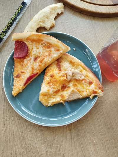 A close-up overhead shot features two slices of pizza on a teal plate, placed on a wooden table. One slice is topped with pepperoni and appears to have some red sauce peeking through the melted cheese. The other slice seems to be topped with chicken and cheese. To the left of the plate is a foil wrapper with text in an East Asian script, likely containing chopsticks. A partially eaten piece of crust rests above the pizza slices. A glass bottle containing a reddish liquid is visible on the right side of the frame. The lighting is bright, suggesting an indoor setting, possibly a restaurant or cafe. The image does not contain any visual information that would allow for the determination of its location in Talas City, Kyrgyzstan.