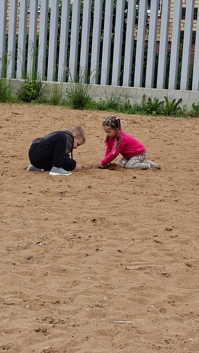 Two children, a boy and a girl, are playing in the sand near a fence in Nytva, Russia. The boy, wearing a dark hoodie and black pants, is crouched on the sand. The girl, in a pink long-sleeved shirt and patterned leggings, is kneeling and digging in the sand with her hands. Both children appear to be focused on their activity. The environment is an outdoor sandy area with a light-colored fence in the background. Green grass and some vegetation are visible behind the fence. The sand is a light brown color and appears to be somewhat damp in places. The overall scene depicts a casual outdoor activity of children playing.
