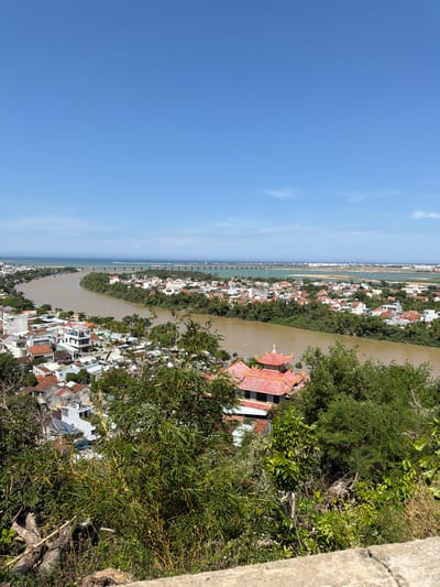 This is an aerial, high-angle shot capturing a panoramic view of a coastal city in Tuy Hoa Ward, Vietnam. The scene is dominated by a wide, brown river winding through a densely populated urban area. Buildings with red-tiled roofs and white facades are clustered along the riverbanks, interspersed with lush green trees and vegetation. In the foreground, a temple-like structure with a distinctive red, tiered roof is visible, partially obscured by dense foliage. Beyond the city, the river flows into the ocean, where a long bridge stretches across the water, connecting different parts of the landscape. The sky is a clear, bright blue with a few wispy clouds. The overall impression is of a vibrant, lived-in city nestled within a beautiful natural environment.