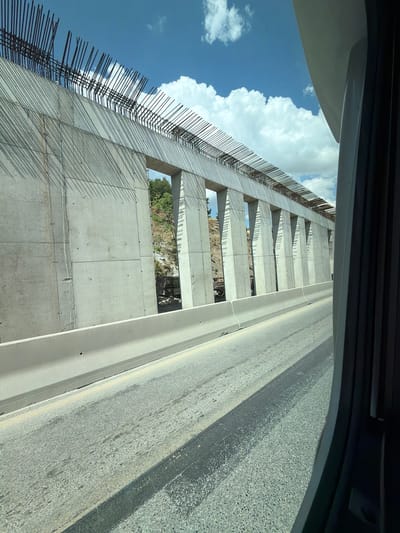 The image shows a partially constructed concrete retaining wall with rebar protruding from the top. The wall is segmented by vertical concrete pillars and has openings that reveal a rocky hillside with vegetation behind it. A concrete barrier runs along the edge of an asphalt road in the foreground, with shadows from the wall and rebar cast across the road. The sky above is blue with white cumulus clouds. The scene is indicative of infrastructure development, possibly a highway or bridge construction, located in the Polichni Municipal Unit, Greece. There are no people visible in the image.