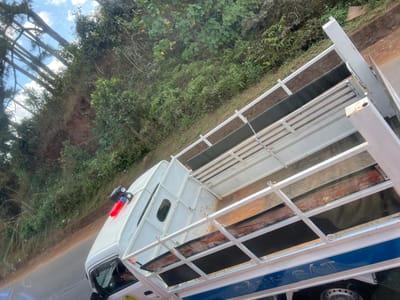An angled view shows the rear of a white truck with a metal frame surrounding the bed. Black material and worn wooden planks line the interior of the truck bed. The cab features a small window and a red and blue light on top. The truck's side displays "CÁNH SÁT" in blue. A road is visible beside the truck, and a lush hillside covered in greenery rises in the background. The sky is blue with some clouds.