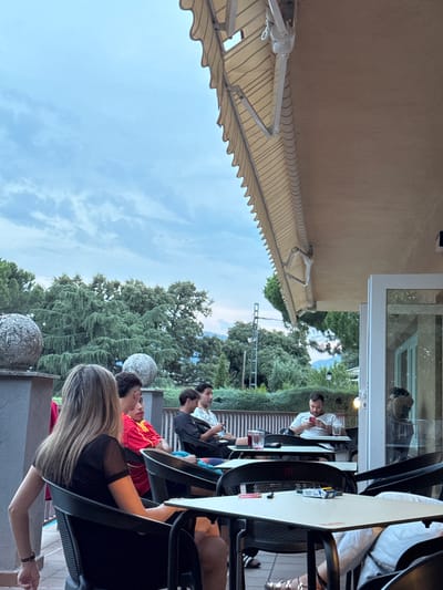 The image depicts an outdoor seating area, possibly a cafe or restaurant, with a group of people seated at tables. The setting appears to be El Escorial, Spain, indicated by the architectural style and the surrounding vegetation which includes mature trees and mountainous terrain in the background, characteristic of the region.
In the foreground, a woman with long blonde hair is seated in a black chair, facing away from the camera, wearing a black top with sheer sleeves. To her right, a young man wearing a red and yellow shirt (possibly a sports jersey) is seated, looking towards his left. Further back, several other individuals are seated at tables. One man is wearing a black shirt and appears to be looking at his phone. Another man, in the background, is seated at a table and looking down at his phone, with a drink in front of him.
The tables are white and have black chairs. On one of the tables, there are a pack of cigarettes and a lighter. The area is covered by a beige awning with a scalloped edge, providing shade. The sky is overcast, suggesting late afternoon or early evening. The environment includes stone decorative spheres on a ledge in the left foreground and a balcony railing in the background. The overall atmosphere is casual and relaxed, with people engaged in their own activities.
