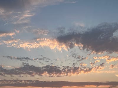 The image shows a wide, unobstructed view of the sky above Rotmanka, Poland, at what appears to be sunset or sunrise. The sky is a gradient of light blue and pale orange, with numerous clouds scattered throughout. Some of the clouds are illuminated by the setting or rising sun, giving them a warm, golden-pink hue, while others are darker and more gray. Wispy, thin clouds are visible higher in the sky, with denser, more textured clouds lower down. There are visible sunbeams or crepuscular rays piercing through gaps in the clouds, particularly on the left side of the image. The horizon line is low and indistinct, suggesting an open landscape or a view from a high vantage point. No people, structures, or distinct land features are visible within the frame, focusing entirely on the atmospheric conditions of the sky.