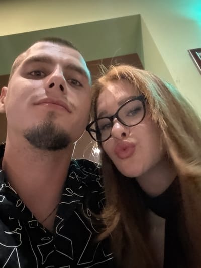 This close-up selfie, taken indoors in Rakitovo, Bulgaria, features a man and a woman. The man, occupying the left side of the frame, has short dark hair, a prominent nose, and a trimmed goatee. He wears a black collared shirt with an abstract white line pattern and a thin necklace. His expression is neutral, and he looks directly into the camera. The woman, positioned on the right, has long, reddish-brown hair and wears black, thick-rimmed cat-eye glasses. Her lips are puckered in a playful "kissing" pose. She is dressed in a black top. The background consists of light green walls, with a corner visible, and a distinct teal-blue light source illuminating the upper right portion of the scene.
