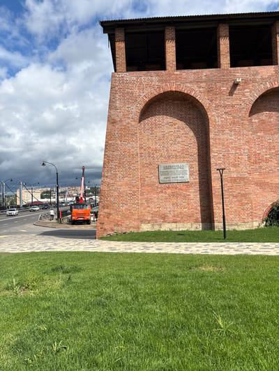 The image shows a close-up view of a section of a large brick wall with arched openings, likely part of a historical fortification or building. The wall is made of red bricks and has a dark wooden roof structure at the top. A plaque is affixed to the wall, bearing text that is difficult to read clearly. To the right of the plaque, a modern, black, minimalist lamppost stands on a patch of green grass.
In the background, a road with several cars is visible, including a white sedan and an orange truck with a crane attached. Streetlights line the road, and a large bridge with a metal structure can be seen in the distance. The sky is partly cloudy with some dark storm clouds gathering.
The scene appears to be an urban environment with a mix of historical architecture and modern infrastructure. The presence of the truck with a crane suggests ongoing construction or maintenance work. The specific location mentioned, Demenshchina, Russia, would imply this is a view of a landmark or historical site within that city.