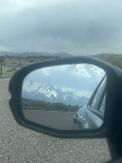 The image depicts a view of a mountain range, possibly the Teton Range, reflected in the side mirror of a vehicle. The mountain range is partially covered in snow and surrounded by a forest. The reflection is framed by the side mirror of the vehicle, and a portion of the vehicle itself is visible. The road is visible on the right side of the mirror. The surrounding environment is hazy or cloudy. The location is likely near Moose, Wyoming, United States of America, given the landscape and potential proximity to the Teton Range.