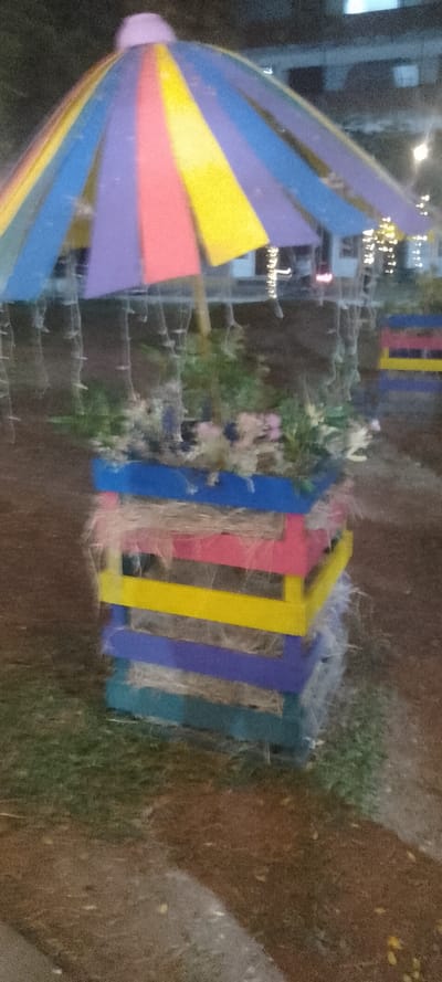 A colorful structure stands in a dark, outdoor setting. It features a tiered box of horizontal planks painted in blue, pink, yellow, and purple, topped with straw. Flowers and greenery are placed in the box. Above, a whimsical, umbrella-like canopy of striped, colorful sections, in shades of purple, blue, yellow, and pink, is suspended. String lights are visible in the background and underneath the umbrella.