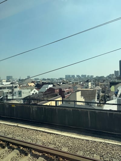 The image depicts an elevated view of a dense urban residential area in Shinagawa, Japan, under a clear, light blue sky. In the immediate foreground, two parallel railway tracks with gravel ballast and concrete sleepers are visible. Directly above the tracks, a dark gray, horizontally oriented concrete or metal barrier spans the lower mid-section of the frame; a small yellow rectangular sign with Japanese text is affixed to its upper left portion.

Beyond this barrier, a densely packed residential neighborhood extends into the midground, composed primarily of multi-story houses and smaller apartment buildings. These structures exhibit a variety of roof styles and colors, including shades of brown, gray, white, red, and yellow. Numerous black utility lines crisscross the midground, running between buildings and poles.

In the distant background, a city skyline features several distinct, taller commercial or residential buildings, appearing somewhat hazy. Three dark, thin overhead wires, likely utility or railway catenary lines, traverse diagonally across the upper portion of the clear blue sky. No individuals, vehicles, or specific actions are discernible within the frame. The scene is illuminated by bright, direct sunlight.