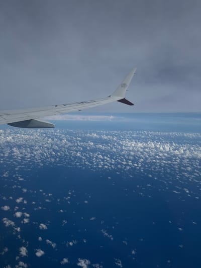 A high-altitude aerial view from the window of a Malaysia Airlines aircraft, visible as a white wing extending from the lower left to the middle right. The wing features the airline's "Wau Bulan" logo and the word "MALAYSIA" on its upper surface, with an engine nacelle visible beneath. Below the wing, a vast expanse of deep blue ocean is observed, dotted with numerous small, scattered white cumulus clouds. A distinct horizon separates the ocean and lower cloud layer from the lighter blue and grey cloudy sky above. No land, structures, or individuals are visible within the frame. The scene is depicted over open water, located in the vicinity of Hallandale Beach, United States.