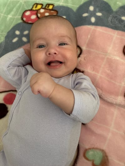The media file shows a close-up, overhead shot of a baby lying on a patterned blanket. The baby, who appears to be a few months old, has fair skin, blue eyes, and a head full of fine, light hair. They are wearing a light purple onesie with a subtle textured pattern. The baby's mouth is open, and they seem to be smiling or laughing, with their tongue visible. Their right hand is raised and clenched into a fist.
The blanket beneath the baby has a colorful, playful design. Parts of the blanket show a pink checkered pattern, a dark blue area with white star-like shapes, and a green area with a pattern that includes brown and red spots resembling mushrooms or polka dots. The lighting suggests it is daytime. The overall scene evokes a sense of warmth, happiness, and domesticity. There is no visible text in the media file. The location context is Wolfsburg, Germany.