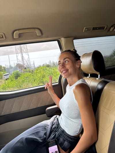 A young adult female is seated in the rear passenger compartment of a vehicle in Paje, Tanzania. She has light skin, dark hair styled in two top buns, and wears a light grey ribbed sleeveless crop top and dark grey sweatpants. Her facial expression is a smile, directed towards her left, and her right hand is raised, forming a peace sign. A black smartphone with an illuminated screen rests on her lap. The vehicle's interior features two-tone beige and light brown patterned seating, a beige headliner, an overhead grab handle, and an air vent. Visible through the window is an exterior scene consisting of dense green foliage and trees, an overcast sky, a prominent metal lattice tower (likely an electricity pylon), and a distant white SUV positioned on an unpaved road.