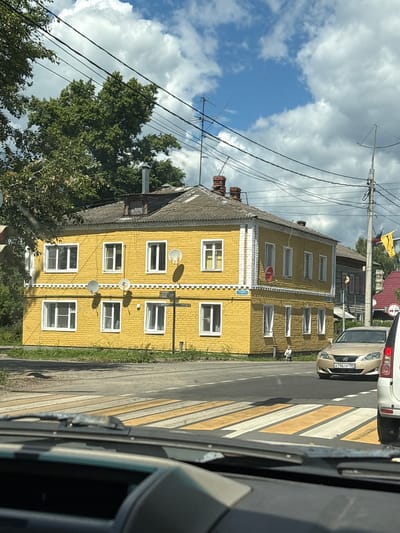 In the city of Pereslavl-Zalessky, Russia, a yellow brick building with a grey roof stands prominently, its facade featuring multiple windows. Several satellite dishes are mounted on the exterior walls. In the foreground, a pedestrian crossing with yellow and white stripes is visible. Two vehicles are present: a gold sedan with a license plate reading "K796TP 799" and a white vehicle partially visible to the right. A street signpost stands near the building. In the background, a leafy tree provides shade, and overhead, electrical wires crisscross under a cloudy blue sky. A small child is seated on the ground near the building.