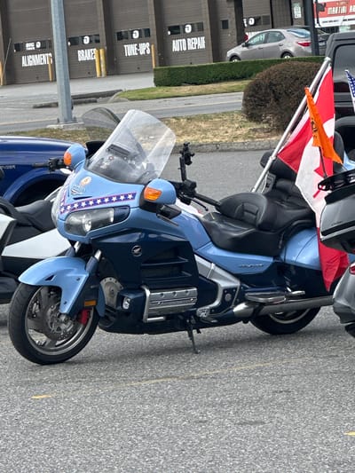 In a parking lot in the Township of Langley, Canada, a light blue Honda Gold Wing motorcycle is parked. The motorcycle is adorned with a Canadian flag and an American flag, as well as a decal on the front fairing featuring stars and stripes. In the background, a row of garage bays are visible with signs indicating "BIG O TIRES," "ALIGNMENTS," "BRAKES," "TUNE-UPS," and "AUTO REPAIR." A silver sedan is parked in front of one of the bays. The scene appears to be an outdoor commercial area.