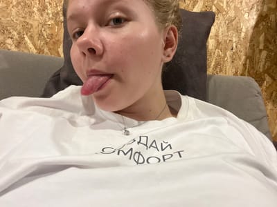 A person is lying down, with their head angled towards the camera. They are wearing a white t-shirt with dark text printed on it. The text is upside down and appears to be in Russian, with the words "ДАЙ СМОТР" visible. The person has their tongue sticking out. Their face is slightly flushed, and they have light-colored hair. A delicate silver necklace with a small charm is visible around their neck. The background consists of a textured wooden surface, possibly OSB board, and a dark gray fabric surface. The lighting suggests an indoor setting. The image orientation is rotated 90 degrees counter-clockwise from standard portrait view.
