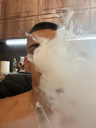 The image shows a close-up of a person exhaling a large plume of white vapor. The person is mostly obscured by the vapor, but their profile is visible, including their ear, neck, and a gold chain. Their hair is short and dark. In the background, there are wooden cabinets and a bright light source. To the right of the person's neck, a silver cylindrical object, possibly a vape device, is visible emitting more vapor. Below the vapor, a black puffer jacket is partially visible. The scene appears to be indoors, possibly a kitchen, given the cabinets and what looks like a bottle and a roll of paper towels on a counter. The lighting suggests it is daytime or an indoor setting with bright artificial lights. The image is taken from a side profile and is rotated so the person's head is to the left. There is no visible text in the image.