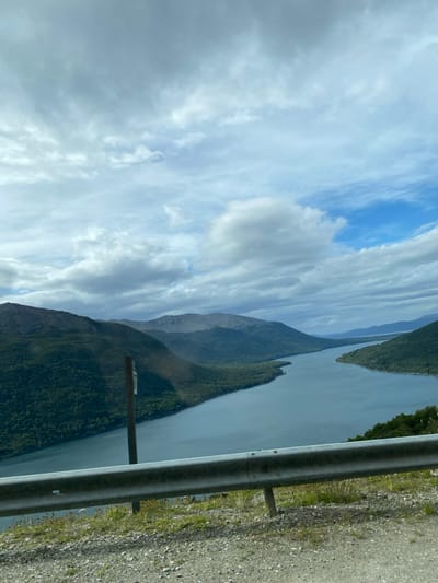 The photograph depicts a scenic fjord landscape under a cloudy sky. A long, narrow body of water is flanked by steep, green mountains. A metal guardrail runs along the right edge of the frame, suggesting a road or viewpoint. The sky is filled with dynamic cloud formations.